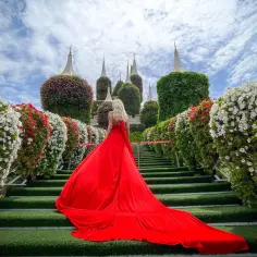 Kobieta w czerwonej sukni na schodach w Miracle Garden w Dubaju, otoczona kwitnącymi krzewami i wieżami z kwiatów.