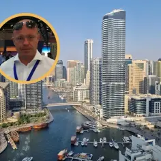 Mężczyzna w okularach na tle nowoczesnych apartamentów w Dubaju. Widok na kanał z łodziami i drapacze chmur.