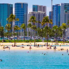Plaża w Honolulu, palmy, turkusowa woda i wysokie budynki. Ludzie odpoczywają, pływają i opalają się. Zastanawiasz się, ile się leci do Honolulu?
