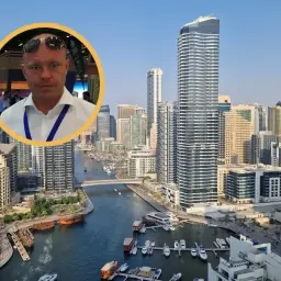 Mężczyzna w okularach na tle nowoczesnych apartamentów w Dubaju. Widok na kanał z łodziami i drapacze chmur.