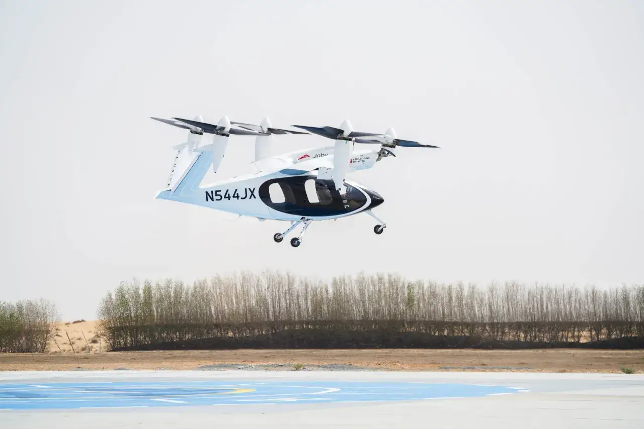 Biała, elektryczna taksówka powietrzna Joby Aviation w locie nad pustynnym krajobrazem. Przyszłość transportu w Dubaju.