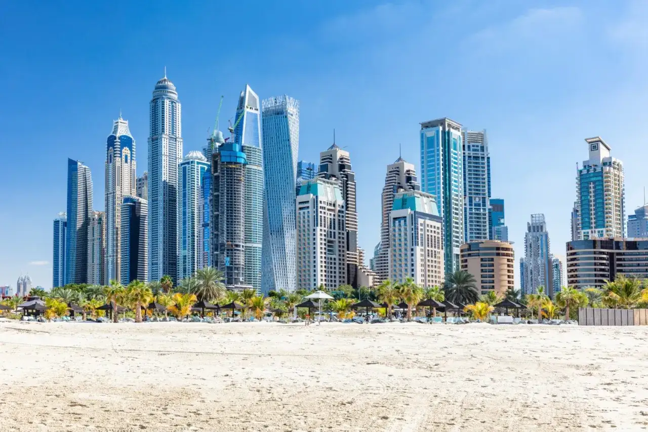 Widok na nowoczesne apartamenty w Dubaju z piaszczystej plaży. Palmy i parasole tworzą wakacyjny klimat.